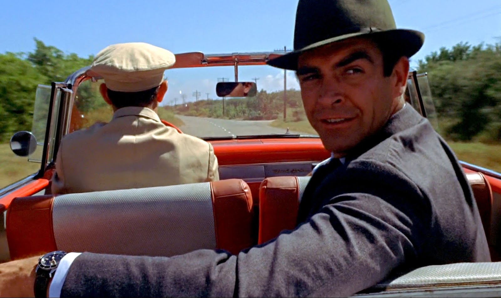 Sean Connery as James Bond sits in the rear seat of a convertible, with his Rolex watch visible.